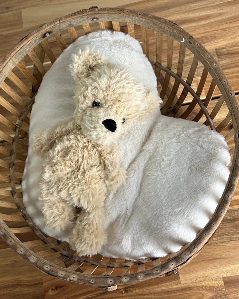 May include: A plush teddy bear with tan fur and black eyes rests on a white, fluffy pillow inside a round, wooden basket. The basket has a woven design and is set against a wooden surface.