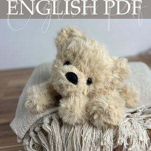 Op de afbeelding: Een zachte, beige teddybeer met een zwarte neus en ogen ligt op een gefranjerde deken. De afbeelding bevat de tekst "Crochet Pattern ENGLISH PDF", wat duidt op een downloadbaar haakpatroon.