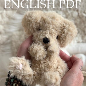 Op de afbeelding: Een gehaakte, beige puppy met een zwarte neus en kleine roze accenten wordt in handen gehouden. De afbeelding bevat de tekst "Crochet Pattern ENGLISH PDF" en de naam van de ontwerper. De puppy is gemaakt van zacht, pluizig materiaal.