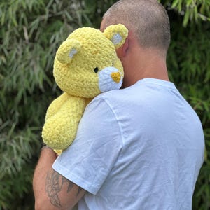 K&ouml;nnte beinhalten: Ein gro&szlig;er, handgefertigter gelber Teddyb&auml;r mit wei&szlig;en Akzenten an Ohren und Schnauze. Das Pl&uuml;schtier ist aus einem weichen, strukturierten Garn gefertigt und hat einen freundlichen Ausdruck. Ein kuscheliges, tr&ouml;stendes Stofftier.