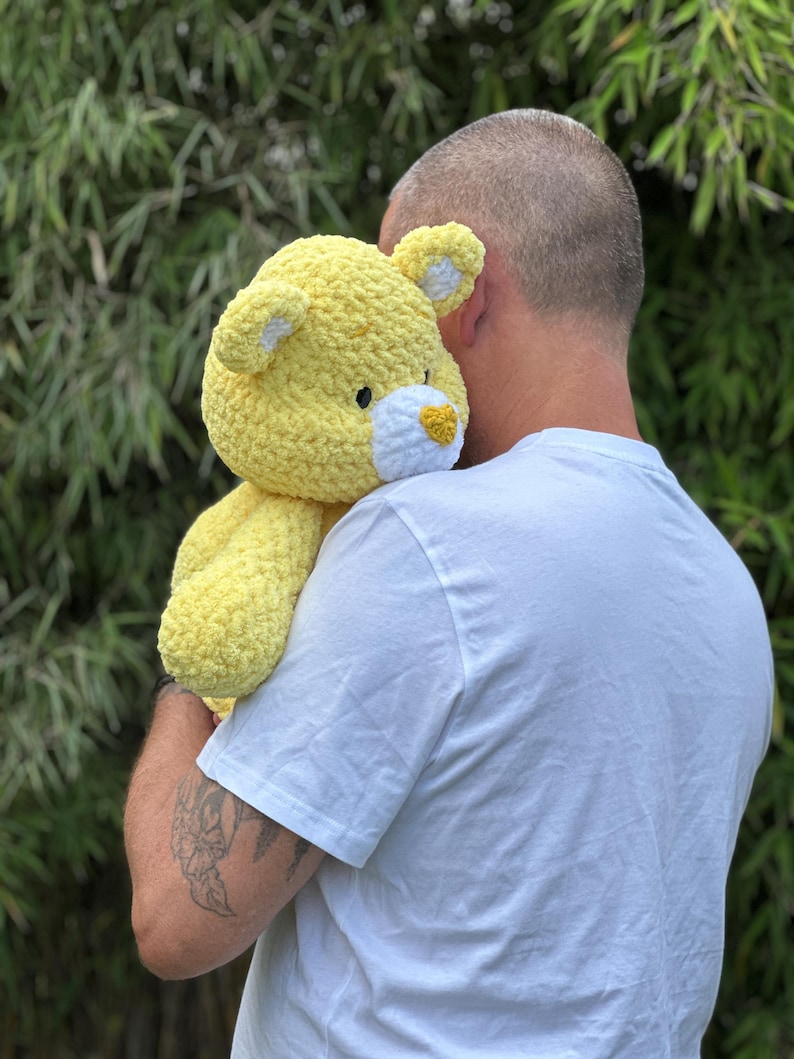K&ouml;nnte beinhalten: Ein gro&szlig;er, handgefertigter gelber Teddyb&auml;r mit wei&szlig;en Akzenten an Ohren und Schnauze. Das Pl&uuml;schtier ist aus einem weichen, strukturierten Garn gefertigt und hat einen freundlichen Ausdruck. Ein kuscheliges, tr&ouml;stendes Stofftier.