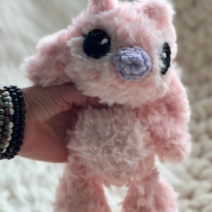 May include: A plush stuffed animal made of soft, fuzzy light pink yarn. The creature features large, round black eyes with small white highlights, a small lavender-colored nose, and two pointed ears. The texture of the yarn is shaggy and dense.