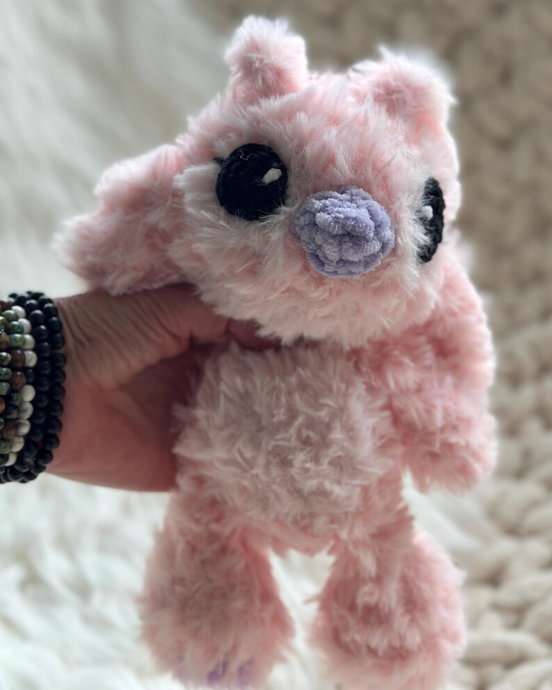 May include: A plush stuffed animal made of soft, fuzzy light pink yarn. The creature features large, round black eyes with small white highlights, a small lavender-colored nose, and two pointed ears. The texture of the yarn is shaggy and dense.