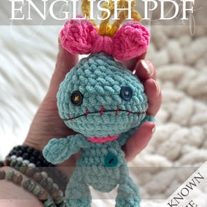 May include: A light blue crochet doll with a large head, a pink bow, and yellow hair. It features two mismatched buttons for eyes, a red stitched mouth, and a small blue button on its chest. The text reads Crochet Pattern ENGLISH PDF.