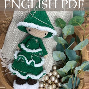May include: A handmade crochet doll wearing a green dress and hat with white trim. The doll has a beige head and brown shoes. The text "ENGLISH PDF" is visible. The doll is displayed in a wooden bowl with decorative elements.
