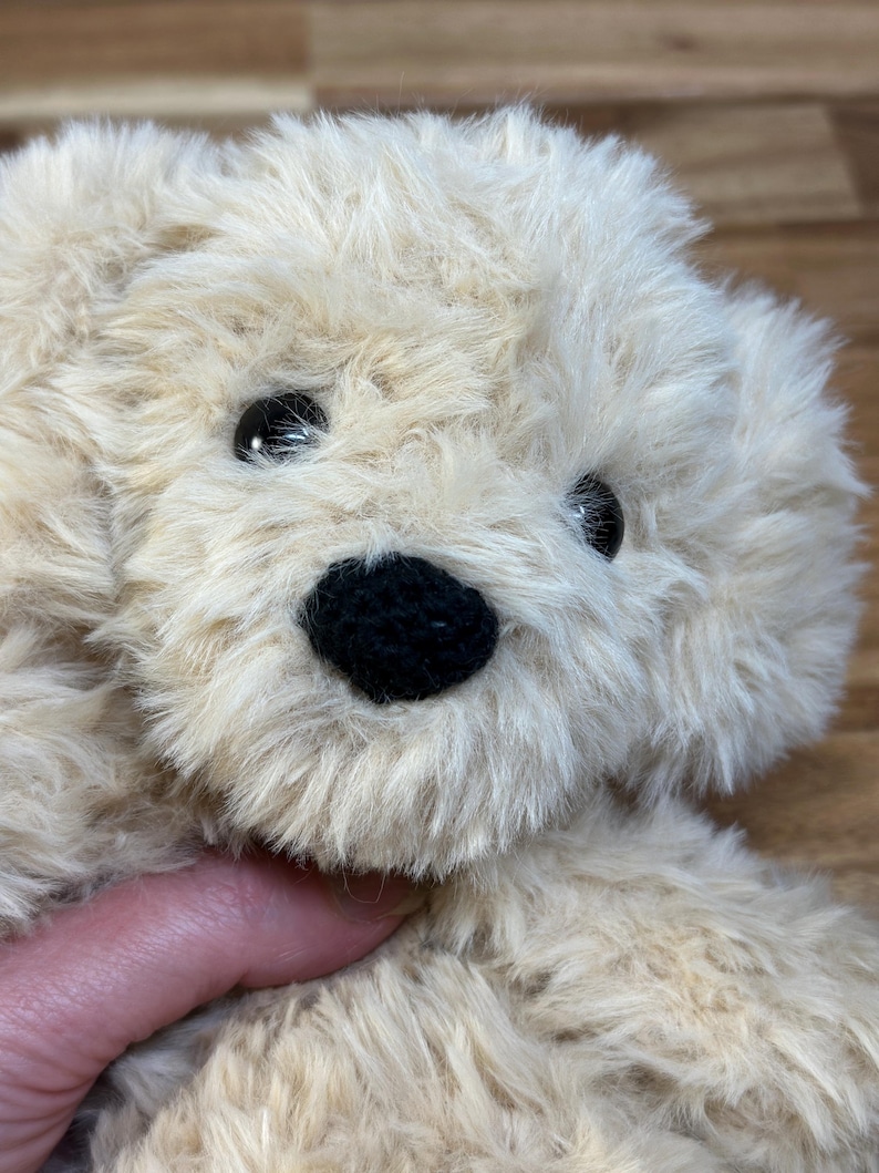 May include: A plush, beige teddy bear with a black nose and black eyes. The soft toy has fluffy fur and is partially visible, suggesting a close-up shot. The bear is a classic children's toy.