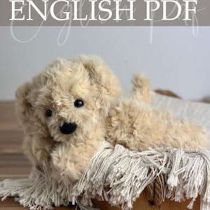 Op de afbeelding: Een zacht, beige gehaakt puppy speelgoed met een zwarte neus en ogen, liggend op een gefranjerde deken. De afbeelding bevat de tekst "Crochet Pattern ENGLISH PDF" en de naam van de ontwerper.