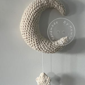 Könnte beinhalten: Ein beigefarbenes, gehäkeltes Halbmond-Mobile mit einem baumelnden Stern. Der Mond hängt an einer weißen Schnur und der Stern ist mit einem Knoten an der Schnur befestigt. Der Mond trägt die Aufschrift "Handmade by Mo".