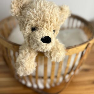 May include: A plush, tan teddy bear with a black nose and eyes peeking over the edge of a wooden basket. The bear has soft, textured fur and a classic teddy bear design. The basket has a light brown color and a woven pattern.