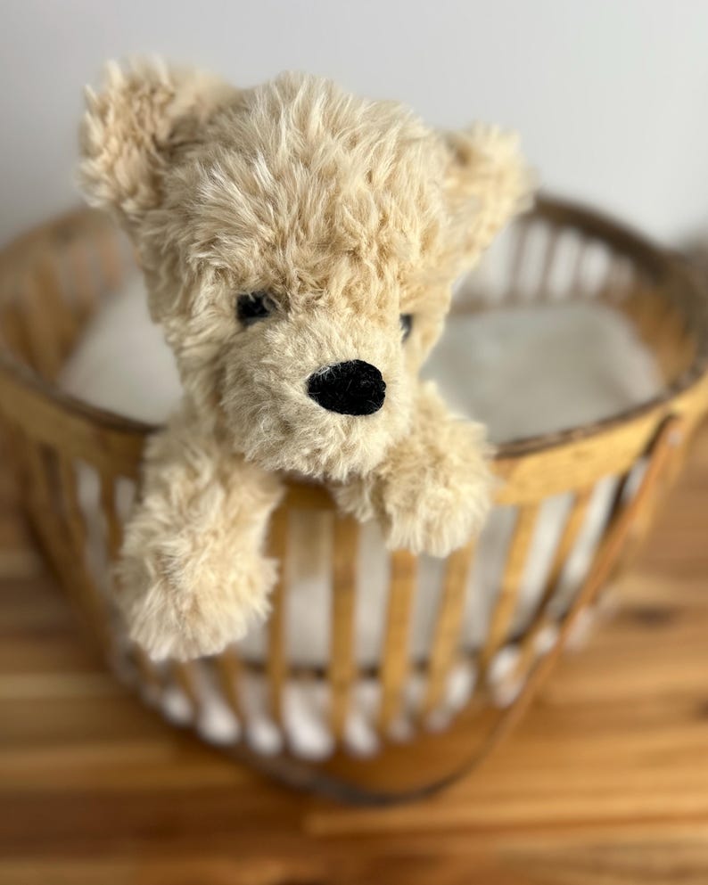 May include: A plush, tan teddy bear with a black nose and eyes peeking over the edge of a wooden basket. The bear has soft, textured fur and a classic teddy bear design. The basket has a light brown color and a woven pattern.