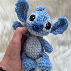 May include: A blue and light blue crocheted plush toy featuring large ears, a rounded head, and a soft textured yarn finish. The character has black embroidered eyes, a dark blue nose, and a light blue belly patch, crafted with a visible stitch pattern.
