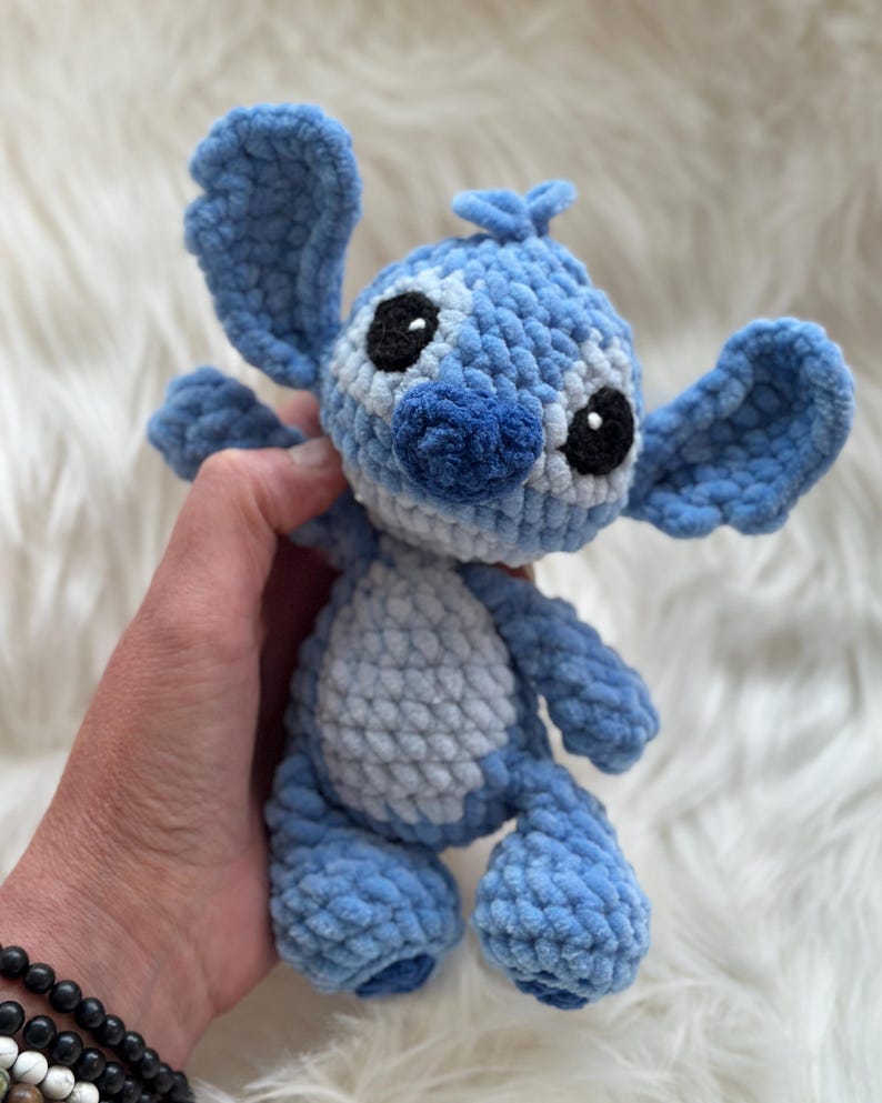 May include: A blue and light blue crocheted plush toy featuring large ears, a rounded head, and a soft textured yarn finish. The character has black embroidered eyes, a dark blue nose, and a light blue belly patch, crafted with a visible stitch pattern.