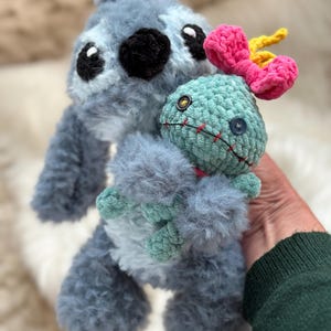 May include: A fuzzy blue crochet plush toy with large black eyes and a black nose, holding a smaller light green crochet doll. The smaller doll features a bright pink bow on its head, two mismatched button eyes, and a stitched red mouth.