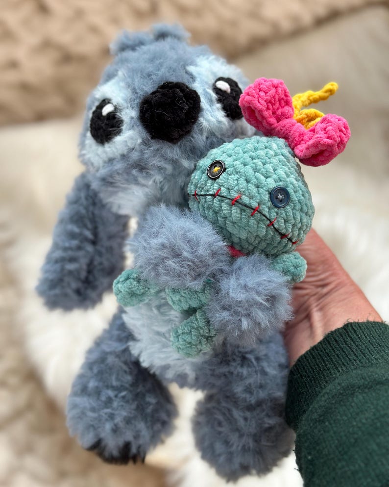 May include: A fuzzy blue crochet plush toy with large black eyes and a black nose, holding a smaller light green crochet doll. The smaller doll features a bright pink bow on its head, two mismatched button eyes, and a stitched red mouth.