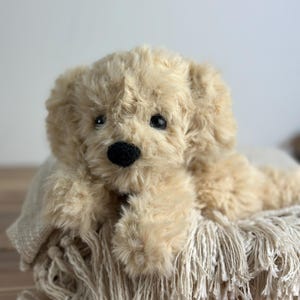 May include: A plush, golden-colored puppy dog toy with a black nose and dark eyes. The stuffed animal is lying on a cream-colored fringed blanket. The toy has soft, fluffy fur and floppy ears.