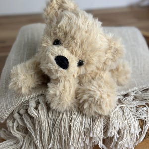 May include: A plush, beige teddy bear with a black nose and eyes rests on a fringed, light-colored blanket. The bear has soft, fluffy fur and is lying down. The blanket has a textured weave and a decorative fringe.