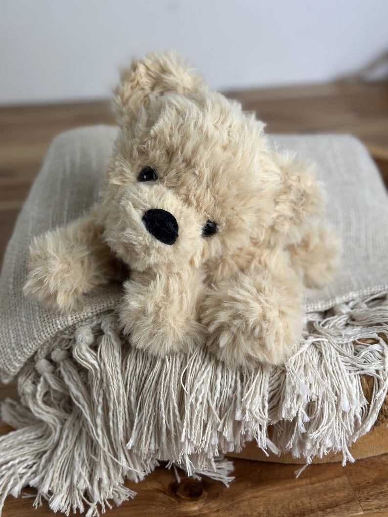 May include: A plush, beige teddy bear with a black nose and eyes rests on a fringed, light-colored blanket. The bear has soft, fluffy fur and is lying down. The blanket has a textured weave and a decorative fringe.