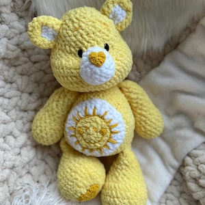 K&ouml;nnte beinhalten: Ein handgefertigter gelber H&auml;kelb&auml;r mit wei&szlig;en Akzenten an Ohren und Brust. Der B&auml;r hat eine wei&szlig;e herzf&ouml;rmige Nase und ein Sonnendesign auf dem Bauch. Das Pl&uuml;schtier ist weich und kuschelig.