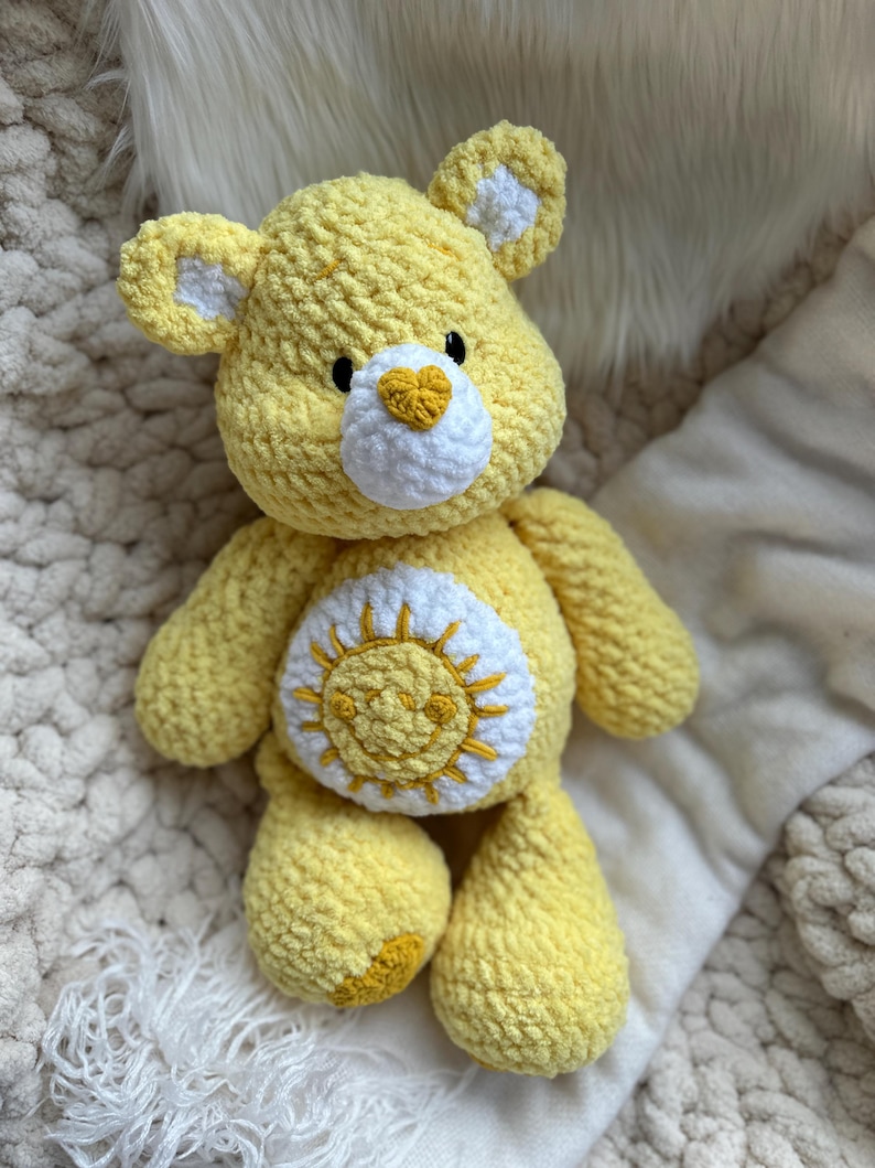 K&ouml;nnte beinhalten: Ein handgefertigter gelber H&auml;kelb&auml;r mit wei&szlig;en Akzenten an Ohren und Brust. Der B&auml;r hat eine wei&szlig;e herzf&ouml;rmige Nase und ein Sonnendesign auf dem Bauch. Das Pl&uuml;schtier ist weich und kuschelig.