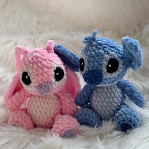 May include: Two handmade crochet plush toys featuring large ears and round black eyes. One plush is light pink with a lighter pink belly and purple nose, while the other is blue with a lighter blue belly and dark blue nose, both made of soft yarn.