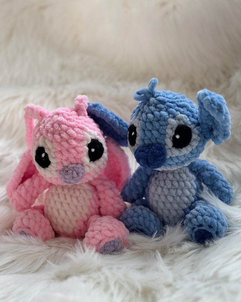 May include: Two handmade crochet plush toys featuring large ears and round black eyes. One plush is light pink with a lighter pink belly and purple nose, while the other is blue with a lighter blue belly and dark blue nose, both made of soft yarn.