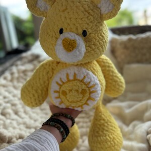 K&ouml;nnte beinhalten: Ein handgefertigter gelber H&auml;kelb&auml;r mit wei&szlig;en Akzenten an Ohren und Brust. Der B&auml;r hat ein l&auml;chelndes Sonnendesign auf dem Bauch und eine herzf&ouml;rmige Nase. Das Kuscheltier ist weich und kuschelig, perfekt f&uuml;r Kinder oder als Dekoration.
