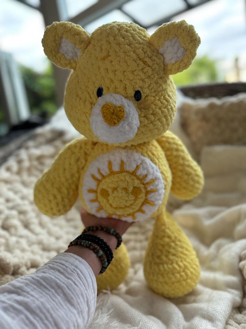 K&ouml;nnte beinhalten: Ein handgefertigter gelber H&auml;kelb&auml;r mit wei&szlig;en Akzenten an Ohren und Brust. Der B&auml;r hat ein l&auml;chelndes Sonnendesign auf dem Bauch und eine herzf&ouml;rmige Nase. Das Kuscheltier ist weich und kuschelig, perfekt f&uuml;r Kinder oder als Dekoration.