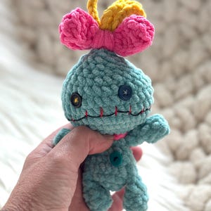 May include: A small, light blue crocheted plush doll with a textured yarn finish. It features a bright pink bow and a yellow tuft on top, mismatched button eyes in black and blue, and a stitched red mouth. A single teal button is on the chest.