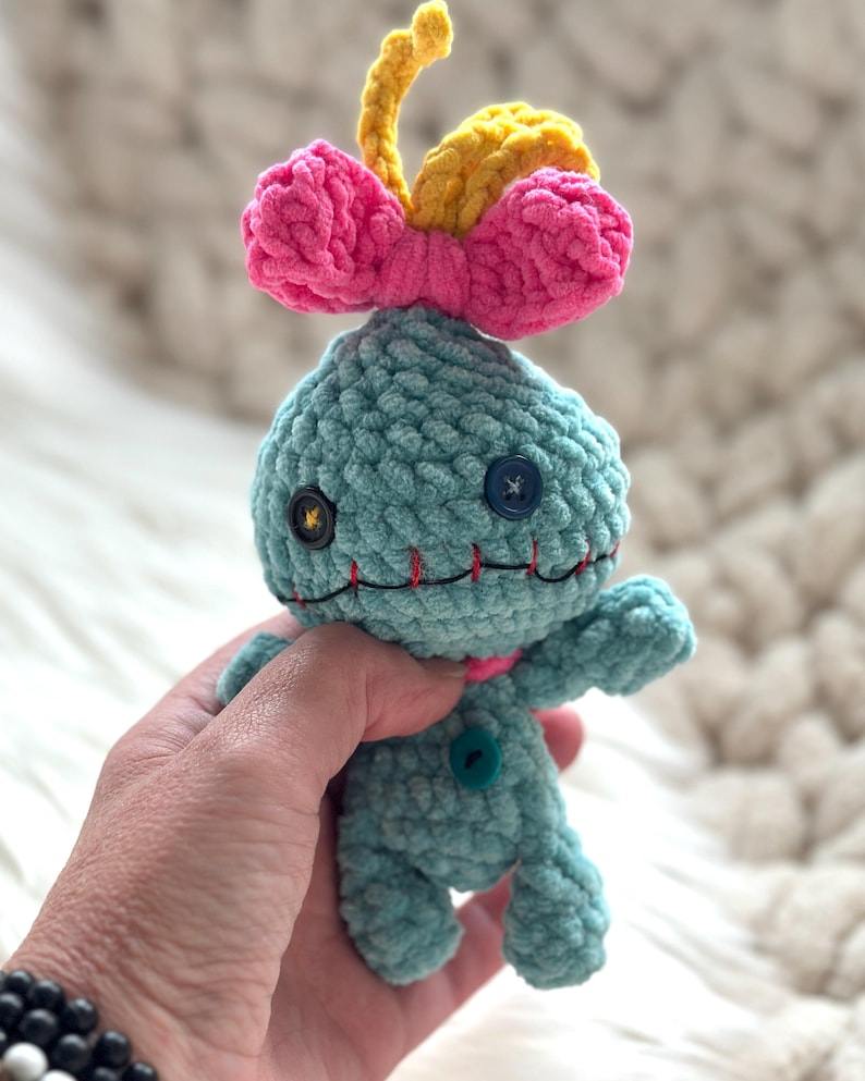 May include: A small, light blue crocheted plush doll with a textured yarn finish. It features a bright pink bow and a yellow tuft on top, mismatched button eyes in black and blue, and a stitched red mouth. A single teal button is on the chest.