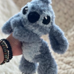 May include: A plush toy character with soft, fuzzy blue fur, large ears, and a light blue belly patch. The creature features black embroidered eyes with small white accents and a black nose, crafted with a textured, shaggy yarn material.