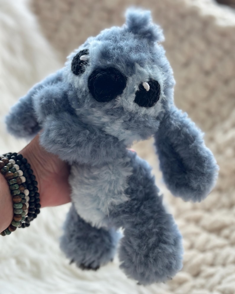 May include: A plush toy character with soft, fuzzy blue fur, large ears, and a light blue belly patch. The creature features black embroidered eyes with small white accents and a black nose, crafted with a textured, shaggy yarn material.
