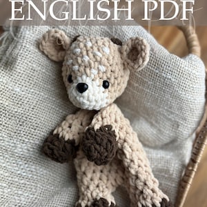 May include: A crocheted deer plushie in tan, cream, and brown. The deer has a cream-colored face with black eyes and a brown nose. The text "Crochet Pattern ENGLISH PDF" is at the top. The plushie is in a woven basket.