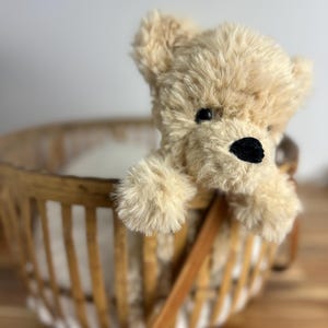 May include: A plush, beige teddy bear with a black nose and eyes peeks out of a woven basket. The basket has a brown leather handle and is made of light-colored wood. The bear's fur is soft and fluffy.