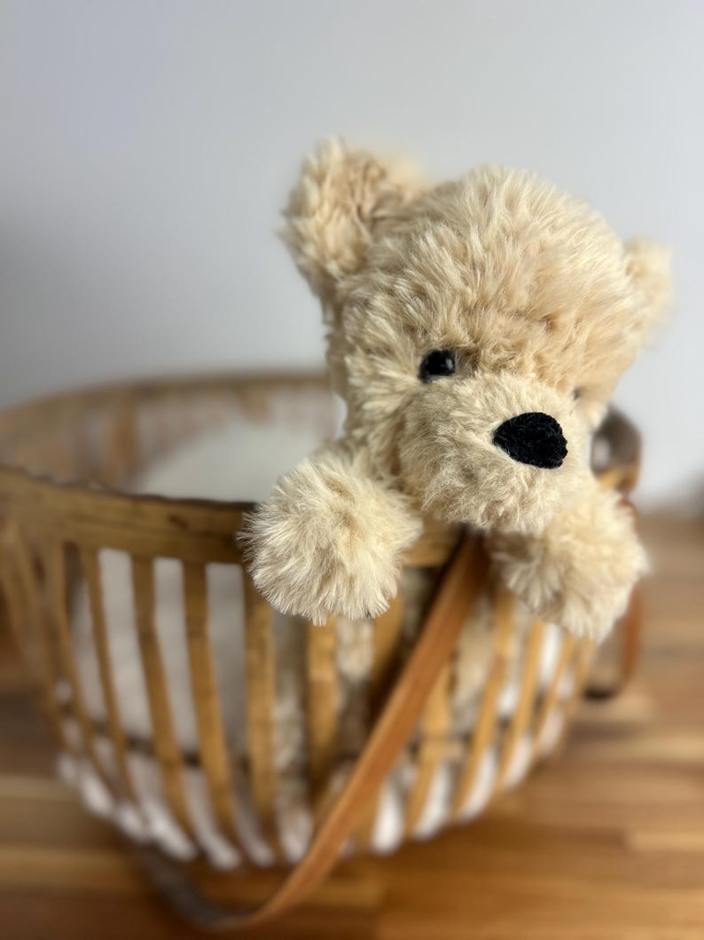 May include: A plush, beige teddy bear with a black nose and eyes peeks out of a woven basket. The basket has a brown leather handle and is made of light-colored wood. The bear's fur is soft and fluffy.