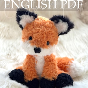 May include: A crocheted fox plush toy with orange, white, and black fur. The fox has a fluffy texture, black eyes, and a white-tipped tail. The text "Crochet Pattern ENGLISH PDF" is displayed at the top of the image.