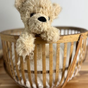 May include: A plush, tan teddy bear with black eyes and nose peeks out of a round, woven bamboo basket. The basket has a handle and is set on a wooden surface. The bear's fur is soft and fluffy.