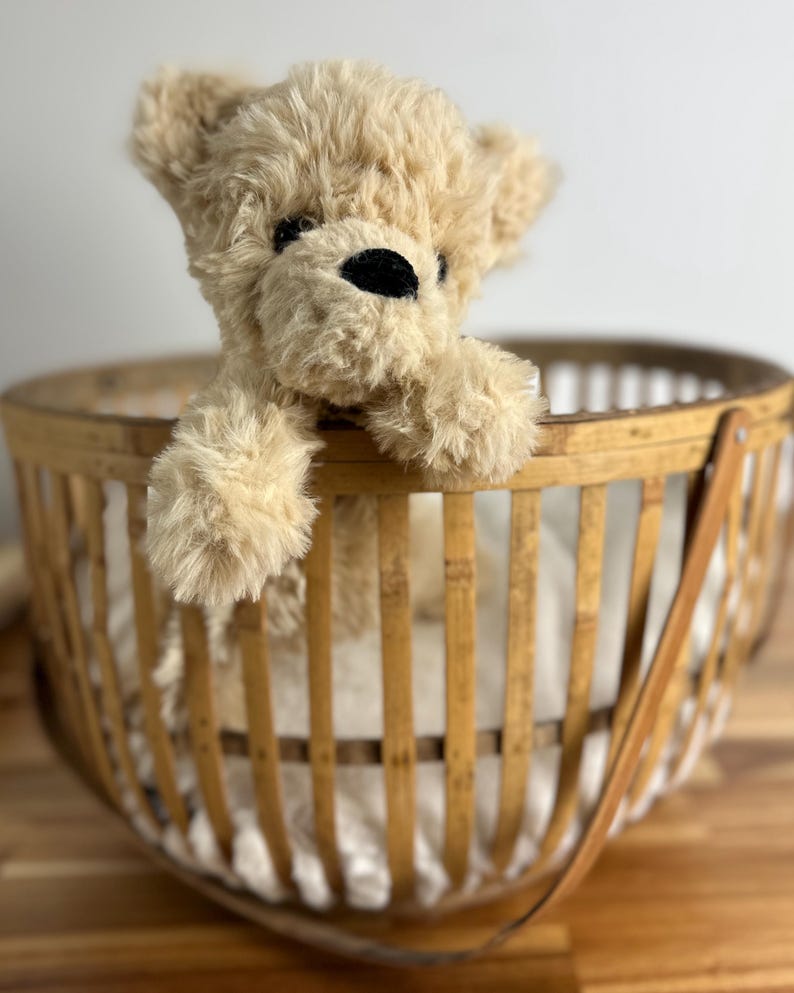 May include: A plush, tan teddy bear with black eyes and nose peeks out of a round, woven bamboo basket. The basket has a handle and is set on a wooden surface. The bear's fur is soft and fluffy.