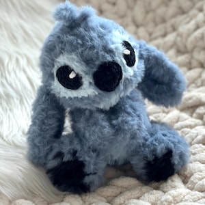 May include: A plush toy crafted from fuzzy blue yarn, featuring large black embroidered eyes with white accents, a black nose, and long ears. The creature has a rounded head, a soft textured body, and dark black patches on its feet, all rendered in a soft, tactile material.