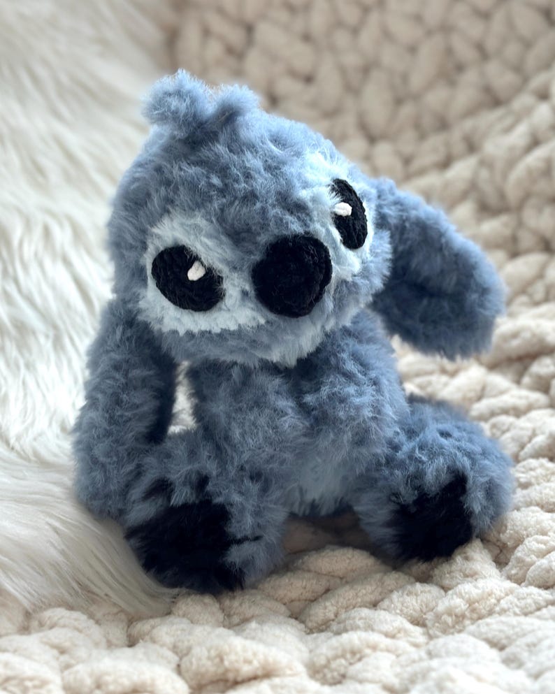 May include: A plush toy crafted from fuzzy blue yarn, featuring large black embroidered eyes with white accents, a black nose, and long ears. The creature has a rounded head, a soft textured body, and dark black patches on its feet, all rendered in a soft, tactile material.