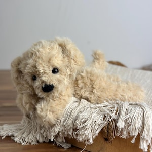 May include: A plush, cream-colored toy dog with a black nose and eyes, lying on a fringed blanket inside a wooden basket. The dog has soft, curly fur and is positioned as if relaxing.