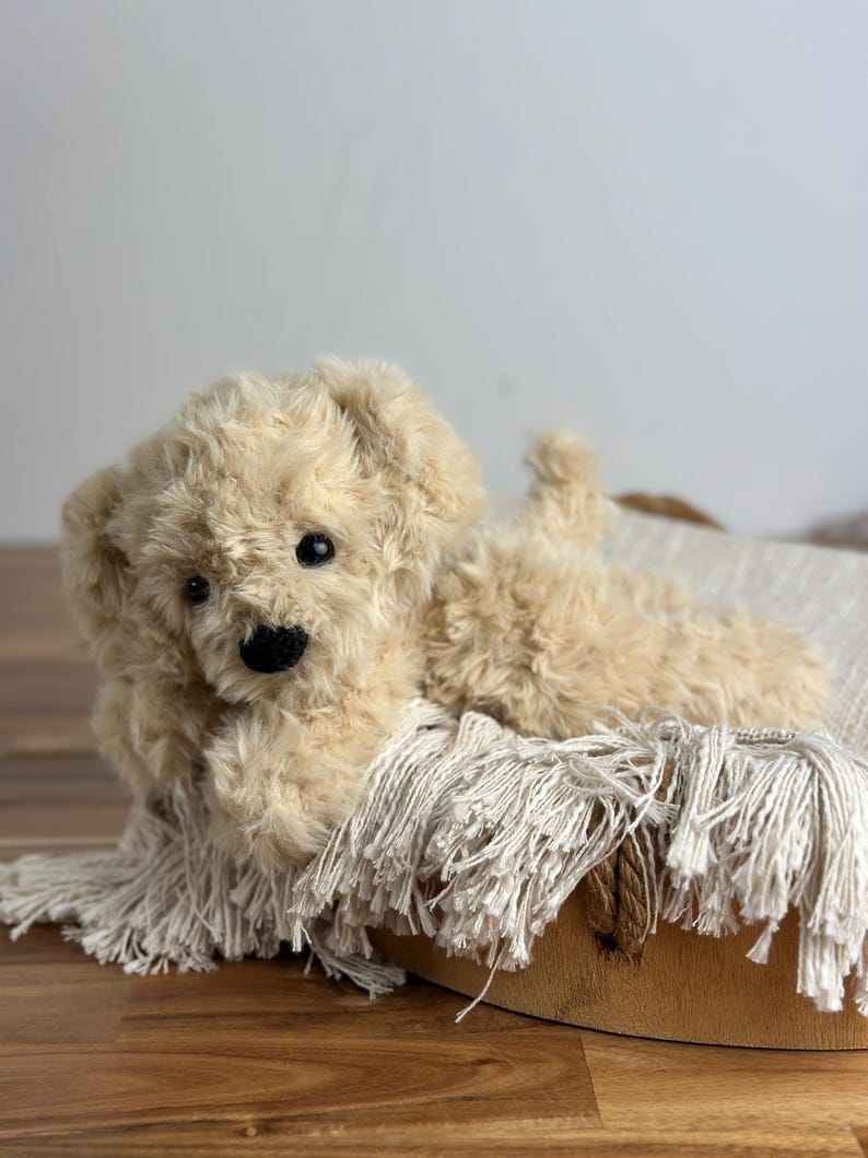 May include: A plush, cream-colored toy dog with a black nose and eyes, lying on a fringed blanket inside a wooden basket. The dog has soft, curly fur and is positioned as if relaxing.