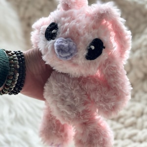 May include: A plush stuffed animal made of soft, fuzzy light pink yarn. The creature features large, dark black oval-shaped eyes and a small, rounded light purple nose. It has long, floppy ears and a textured, shaggy appearance throughout its body.