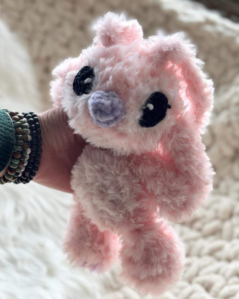 May include: A plush stuffed animal made of soft, fuzzy light pink yarn. The creature features large, dark black oval-shaped eyes and a small, rounded light purple nose. It has long, floppy ears and a textured, shaggy appearance throughout its body.