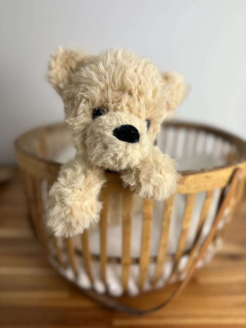 May include: A plush, light brown teddy bear with a black nose and eyes peeking over the edge of a woven basket. The basket has a light brown wooden frame and a white interior.