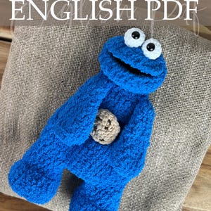 May include: A handmade blue crochet Cookie Monster doll holding a tan crochet cookie. The doll has large white eyes with black pupils. The words "Crochet Pattern ENGLISH PDF" are written at the top of the image.
