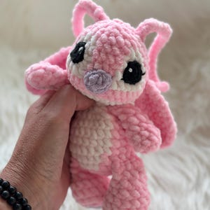 May include: A plush crochet toy featuring a pink and white creature with large ears, black eyes, and a small purple nose. The stuffed animal is crafted from soft, textured yarn and displays a rounded head, a lighter pink belly, and short limbs.
