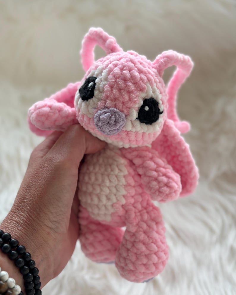 May include: A plush crochet toy featuring a pink and white creature with large ears, black eyes, and a small purple nose. The stuffed animal is crafted from soft, textured yarn and displays a rounded head, a lighter pink belly, and short limbs.