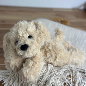 May include: A plush, beige-colored stuffed dog with a black nose and eyes, lying on a fringed, off-white blanket. The dog has soft, fluffy fur and is positioned on a wooden surface.