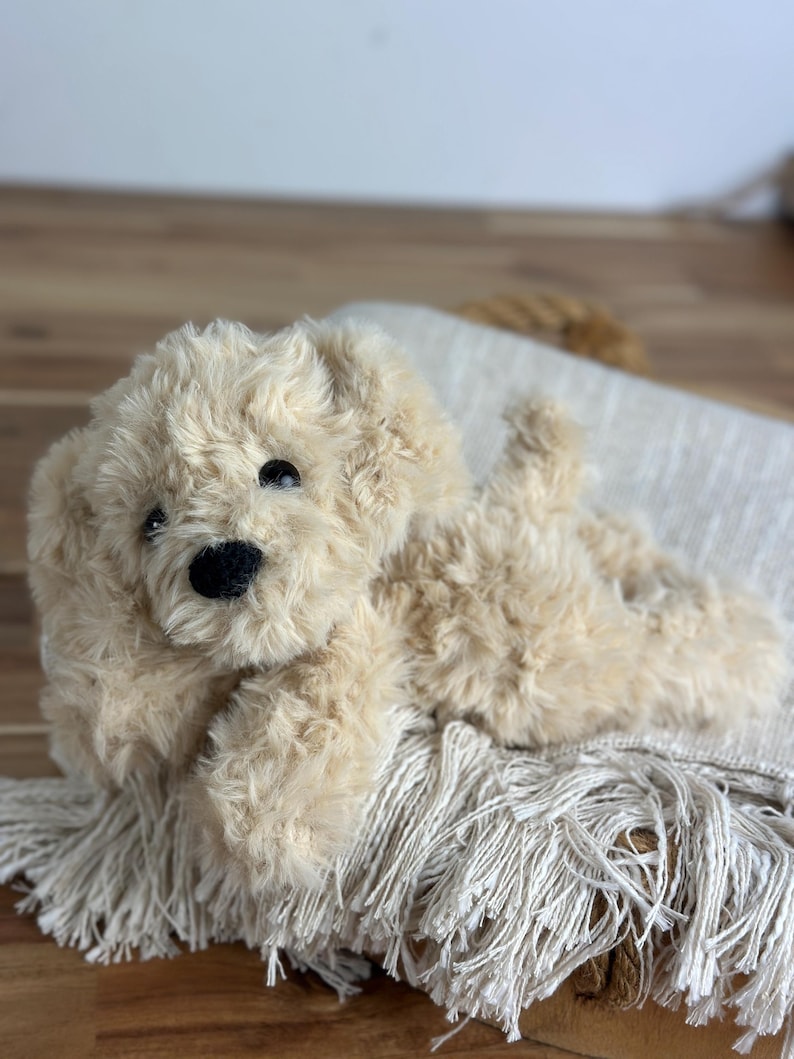 May include: A plush, beige-colored stuffed dog with a black nose and eyes, lying on a fringed, off-white blanket. The dog has soft, fluffy fur and is positioned on a wooden surface.