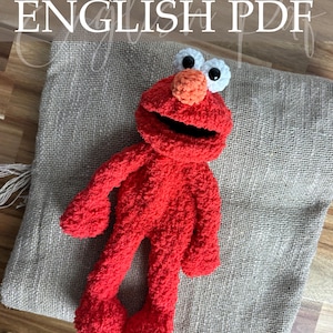 Op de afbeelding: Een rode gehaakte Elmo-pop met witte ogen en een oranje neus, liggend op een jute zak. De afbeelding bevat de tekst "Crochet Pattern ENGLISH PDF" in witte letters. De naam van de ontwerper staat onderaan de afbeelding.